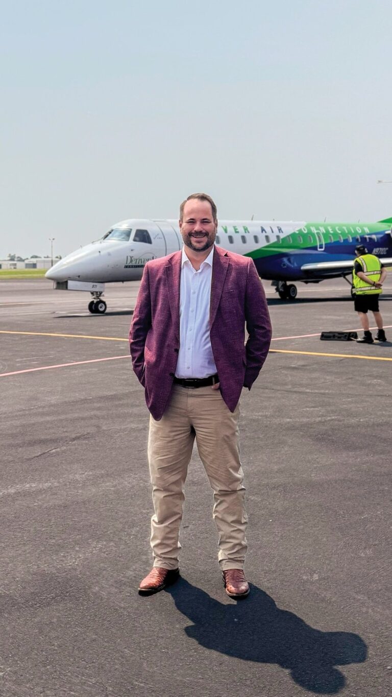 Man in white dress shirt, maroon blazer, and khaki pants standing in front of a small passenger jet on a tarmac